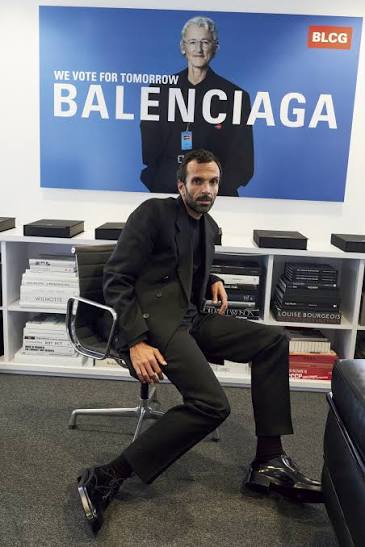Cédric Charbit seated on a chair in an office during his tenure at Balenciaga, with a Balenciaga campaign poster and shelves of books behind him