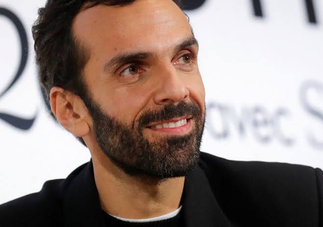 Cédric Charbit photographed smiling at a press appearance in front of a light backdrop