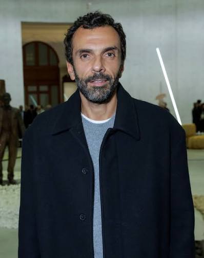 Cédric Charbit standing in a navy overcoat inside a large exhibition space with sculptures visible behind him